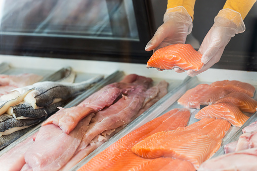 Fresh fish fillets in a display case, for sale in seafood store. We see the hands of a worker, wearing gloves, taking a piece of salmon fillet out of the case.