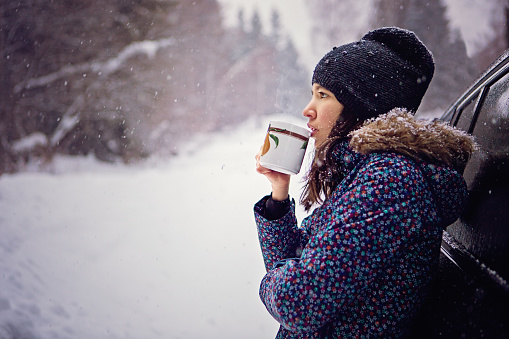 Girl is drinking hot tea in the snowy forest outside the car
