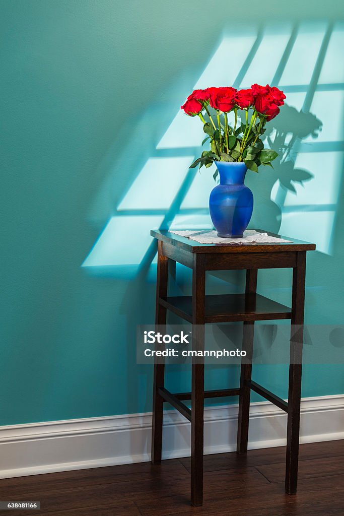 Window light shining on a dozen red roses on table A shaft of sun light coming from a window shining down on a dozen red roses in a blue glass vase, sitting on a vintage oak stand against a blue interior wall. A nice Valentine's Day or special occasion gift to come home to Anniversary Stock Photo