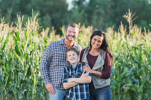 A family with a teenage son standing next to a field of crops. The corn is almost ready for harvesting. The boy is standing in the middle, between his parents. They are looking at the camera, smiling.