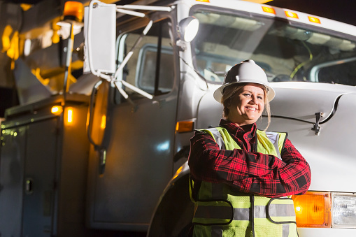 A female worker wearing a hardhat and safety vest standing in front of a work truck. She is a utility worker, engineer or technician.