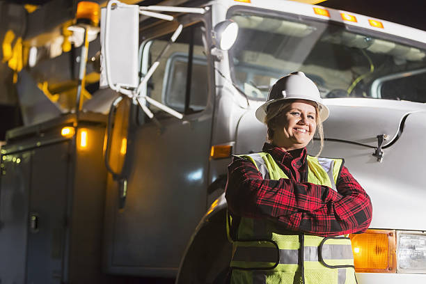 Mature woman in safety vest, hardhat with truck A female worker wearing a hardhat and safety vest standing in front of a work truck. She is a utility worker, engineer or technician. role reversal stock pictures, royalty-free photos & images