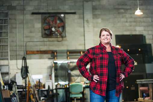 A mature woman in her 50s standing with hands on hips, a worker in a factory warehouse. She is wearing a plaid shirt and jeans. A desk, worktable, equipment and supplies are out of focus in the background.
