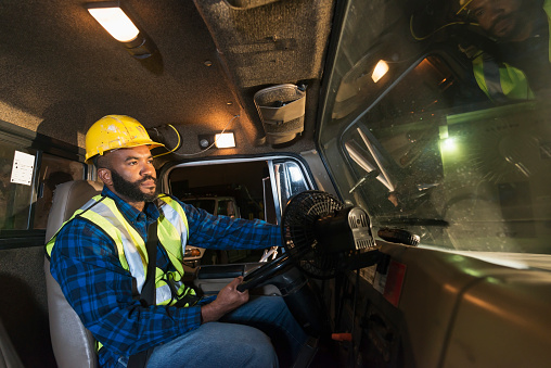 An Afircan American man driving a truck, wearing a hard hat and safety vest.