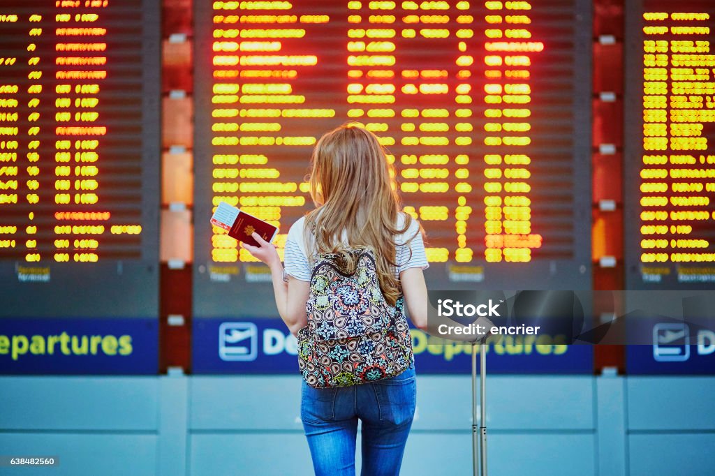 Tourist girl with backpack in international airport Beautiful young tourist girl with backpack and carry on luggage in international airport, near flight information board Airport Stock Photo