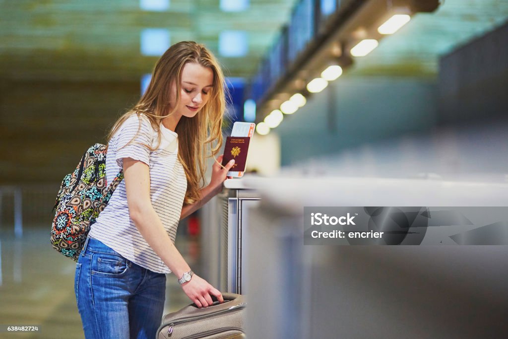 Traveler with backpack in international airport at check-in counter Beautiful young tourist girl with backpack and carry on luggage in international airport at check-in counter, giving her passport to an officer and waiting for her boarding pass Teenager Stock Photo