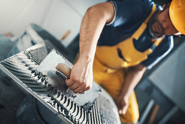 Tile handyman applying adhesive on a tile. Closeup of a handyman applying adhesive with a trowel material onto a tile before installing it on a bathroom wall. He's wearing yellow uniform and helmet. Tilt shot, selective focus. solvent stock pictures, royalty-free photos & images