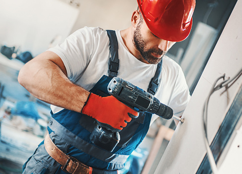 Closeup front view of construction worker placing a plaster board over a wall and using electric screwdriver to tighten it. The worker is wearing blue uniform, red protective gloves and helmet. He's partially unrecognizable with brown beard and mustache.