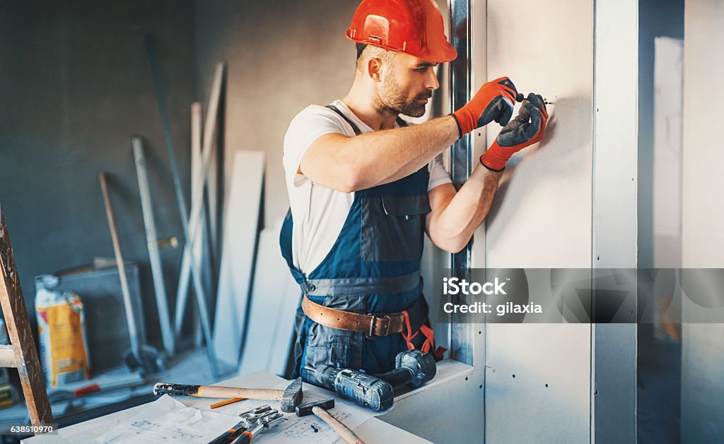 Construction worker routine. Closeup front view of construction worker placing a plaster board over a wall and using electric screwdriver to tighten it. The worker is wearing blue uniform, red protective gloves and helmet. He's partially unrecognizable with brown beard and mustache. Drywall Stock Photo