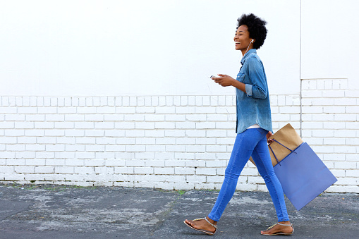 Side portrait of a smiling young woman walking with shopping bags and listening to music on smart phone