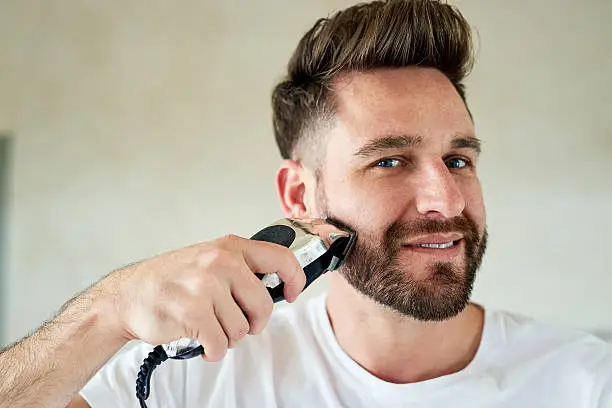 Portrait of a young man shaving his face with an electric razor at home