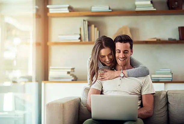 Shot of a young woman hugging her husband while he uses a laptop on the sofa at home