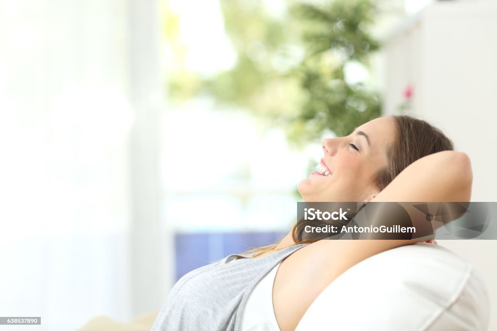 Relaxed girl laughing on a couch at home Side view portrait of a relaxed person laughing sitting on a couch in the living room at home with a window and a green background Under the Arm Stock Photo