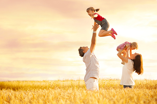 Young playful father is playing with daughter in the field  by throwing up a happy girl in the air and catching her while mother is holding a baby girl.
