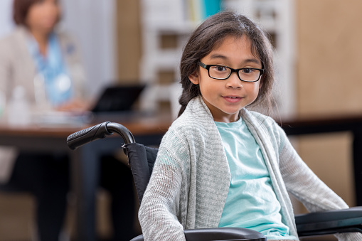 Pretty Filipino elementary age girl in wheelchair waits in doctor's office waiting room.