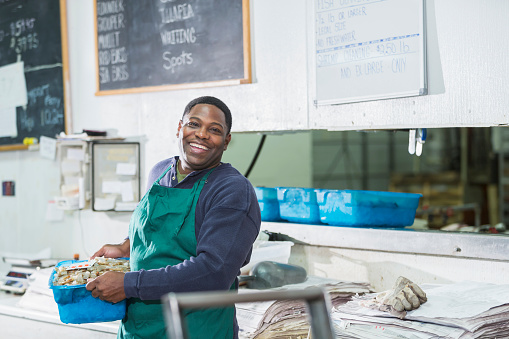 A mature, black man in his 40s working in a seafood market, carrying a tray of fresh, raw shrimp. He is the owner of this small retail business, or a hardworking sales clerk.