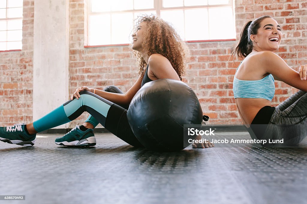 Young women taking a break from workout Happy young female friends sitting on gym floor with medicine ball and smiling. Two young women taking a break from workout. Exercising Stock Photo