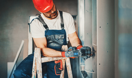 Closeup front view of partially unrecognizable construction worker assembling plaster plates over a wall during a process of home redecoration. He's climbed a wooden ladder and using electric drill with a screwdriver attachment. Wearing blue uniform and red gloves and helmet.