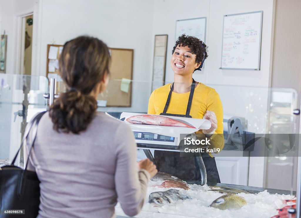 Woman in seafood market helping customer A mixed race woman working in a seafood market, helping a female customer who is buying a fish. She is talking to the customer as she weighs the snapper on a scale. Fish Stock Photo