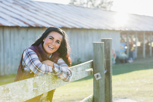 A mature woman in her 40s leaning on a wooden fence outside a farm building on a sunny day, smiling at the camera with her arms folded.