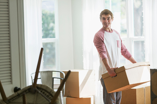 A young man moving into a house or apartment, carrying a cardboard box indoors by a sunny window.