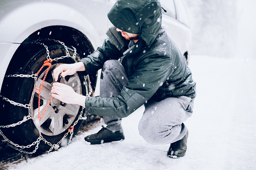 A low angle view of a front wheel drive vehicle on a snowy and ice covered road, a man putting chains on the wheels to improve traction in the winter weather conditions.  Horizontal image with copy space.