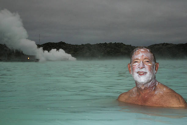 Smiling adult Caucasian male in 60s with silica mud on his face soaks in a milky green geothermal hot spring with steam rising in the distance, Iceland, Europe