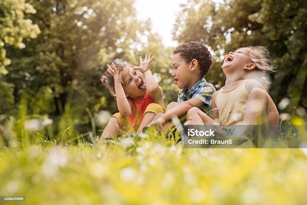 Small friends having fun while relaxing in grass. Three cute children sitting in grass at the park. One of them is laughing while two of them are sticking their tongues out. Child Stock Photo
