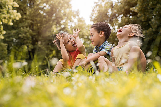 Small friends having fun while relaxing in grass. Three cute children sitting in grass at the park. One of them is laughing while two of them are sticking their tongues out. children only stock pictures, royalty-free photos & images