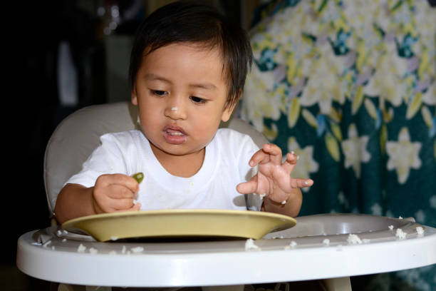 One 1 year old Asian baby boy learning to eat by himself, messy on baby dining chair at home