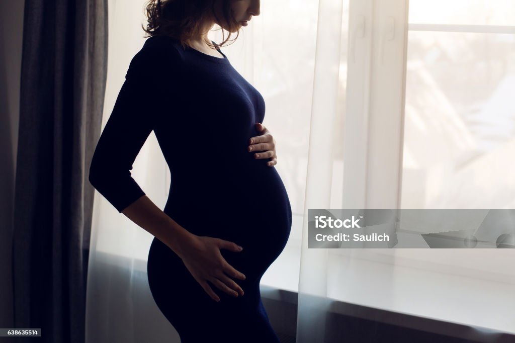 young pregnant girl in a blue bodycon dress standing young pregnant girl in a blue bodycon dress standing at the window of the house Pregnant Stock Photo