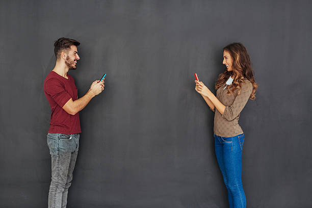 Couple texting in front of empty blackboard Boy and girl holding smart phones standing face to face in front of empty blackboard. face to face stock pictures, royalty-free photos & images