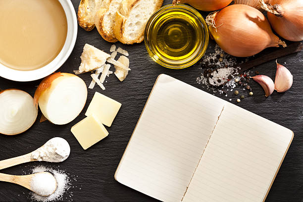 Top view of a dark table with onion soup ingredients and a blank cookbook. At the upper-left corner is visible a bowl filled with beef broth while the other ingredients like onion, bread, garlic, butter, cheese, salt, flour, sugar and olive oil complete de frame. DSRL studio photo taken with Canon EOS 5D Mk II and Canon EF 100mm f/2.8L Macro IS USM