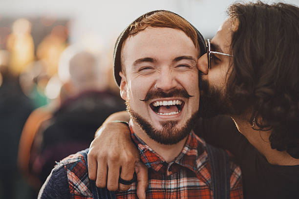 Festival Kisses Young man is recieving a kiss on the cheek by his same sex partner while at a music festival. He is laughing and looking at the camera. man gay stock pictures, royalty-free photos & images