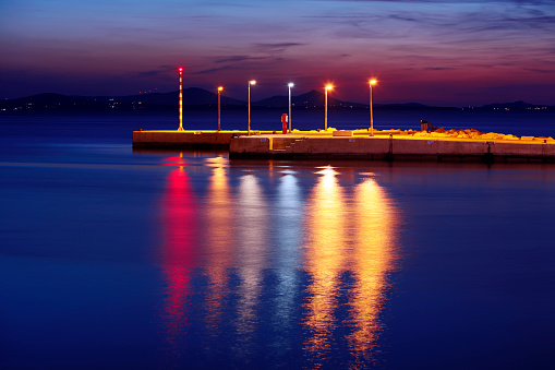 A concept image, a row of colorful lights on the wharf at night indicate direction to the safe harbor, reflections of the lights are visible on the surface of the sea. This image can symbolize many ideas, for example: safe haven, glimmer of hope, light in the darkness, light in the night, safety of navigation, trust, light at the end of the tunnel, 50 Megapixel horizontal image, long exposure with tripod, Naxos island in Greece.