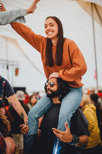 Young adult couple are dancing in a marquee at a music festival. The woman is on her partner's shoulders and has one hand in the air.