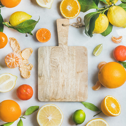Variety of fresh citrus fruit for making juice or smoothie and wooden chopping board over light grey marble background, top view, copy space, square crop. Healthy eating, vitamin, clean eating concept