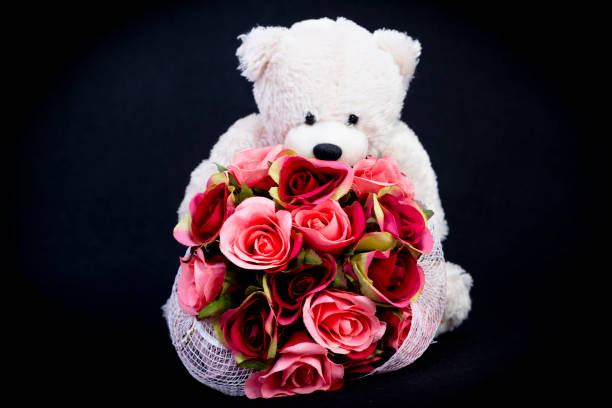 Selective focus at bouquet of beautiful artificial red roses in the arms of blurry white bear doll  on black background, concept love for valentine day or spacial  anniversary