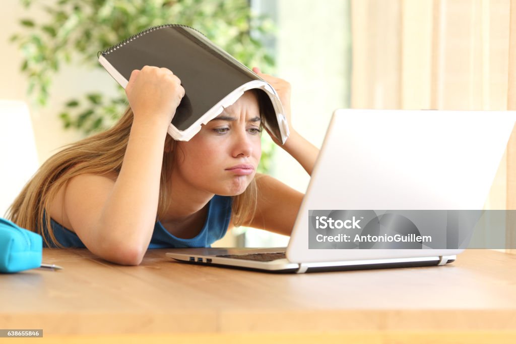 Bored student doing homework Bored student doing homework on line with a laptop blowing with a notebook on the head in a table at home Confusion Stock Photo