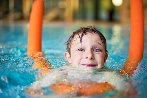 Happy kid learning to swim in indoors swimming pool. The boy is aged 5 and is using orange pool noodle. He is smiling into the camera.