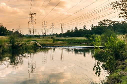 Electricity pylons at sunset with the reflection on river going over a park in Johannesburg, South Africa. Trees and bush along the riverside.