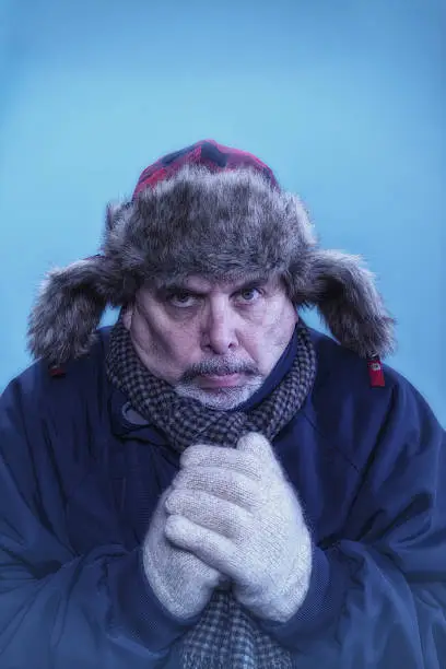 A middle aged 50ish Caucasian male with his hands clasped, all bundled up in a fur trapper hat, scarf, parka and knit gloves trying to stay warm on a very cold day in Winter