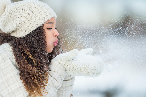 A teenage girl is standing outside in the winter snow and is blowing snow off her gloves.