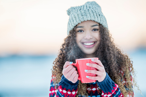 A teenage girl is standing outside in the winter snow and is drinking a cup of hot chocolate.