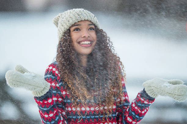 close-up portrait of young smiling caucasian girl in sunny day in winter snowy forest in white cap, beige fur jacket and mittens, blowing on snow in hands, snowflakes flying