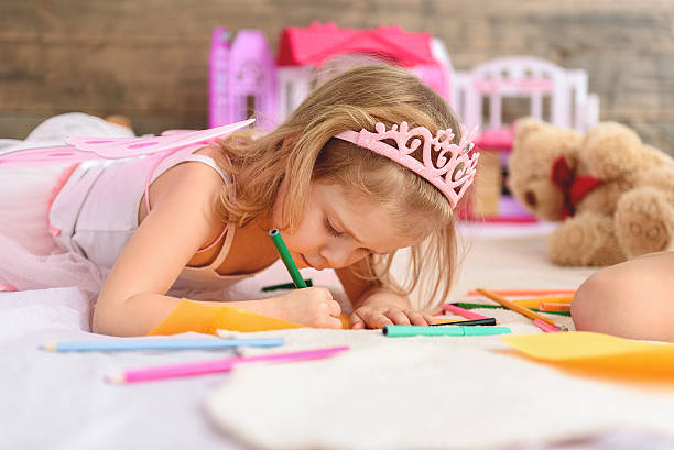 Small kid is trying to paint image with concentration. She is lying on bed. Many colored highlighters are around the girl