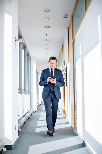 Full length shot of entrepreneur walking through office corridor and reading text message on smart phone