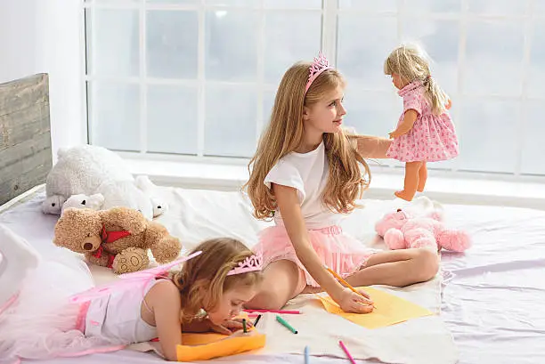 Future artists. Good-looking girls drawing different pictures. Elder sister is painting her doll and smiling. Toys are on the big bed