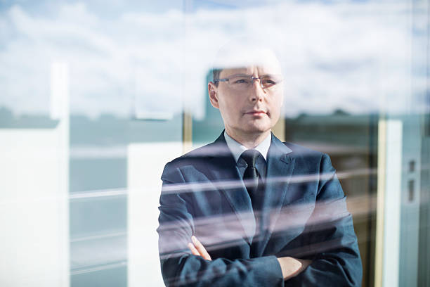 Shot of mature businessman standing behind glass window with arms crossed and thinking