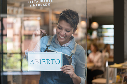 Happy Latin American business owner holding an open sign in Spanish at a restaurant - small business concepts